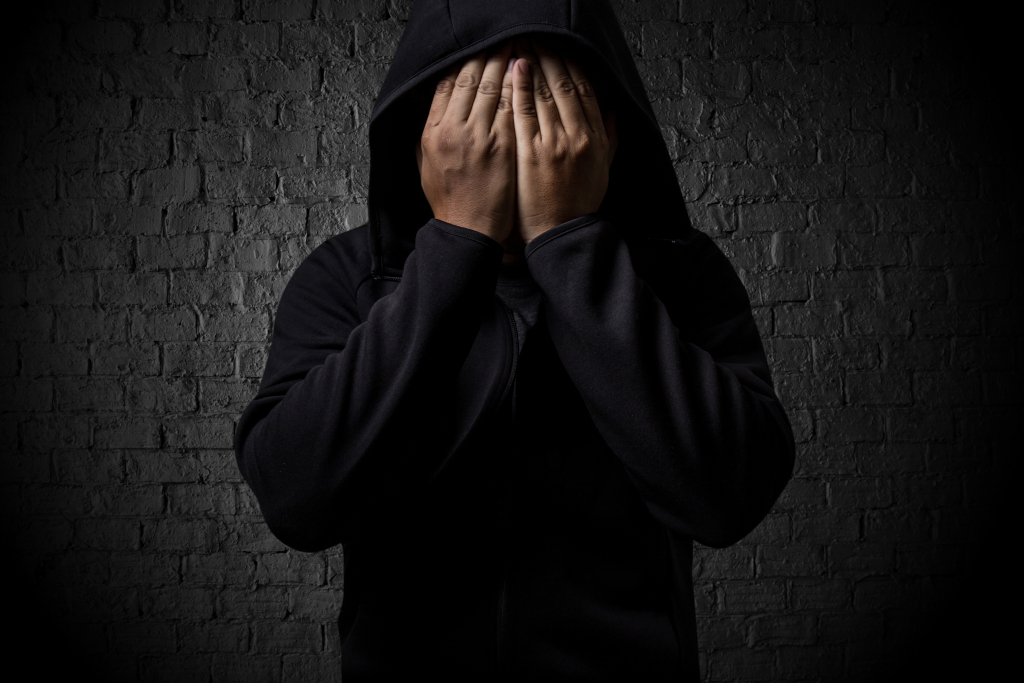A distressed man in a hoodie covers his face with both hands